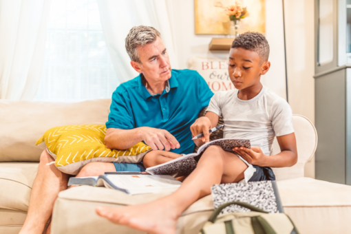 A middle-aged man and a small boy are sitting on a sofa. The man is helping the boy with some homework.