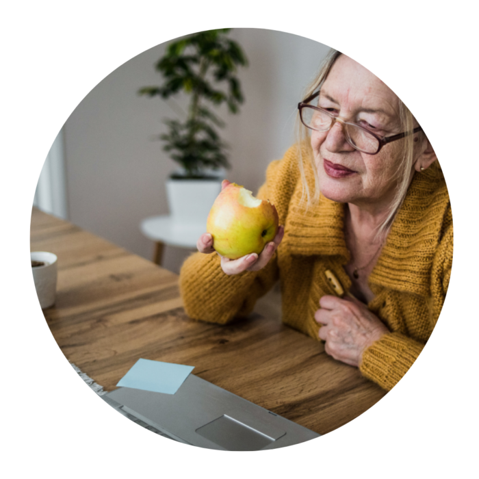 An older woman sitting at the kitchen table eating an apple and looking at her laptop.