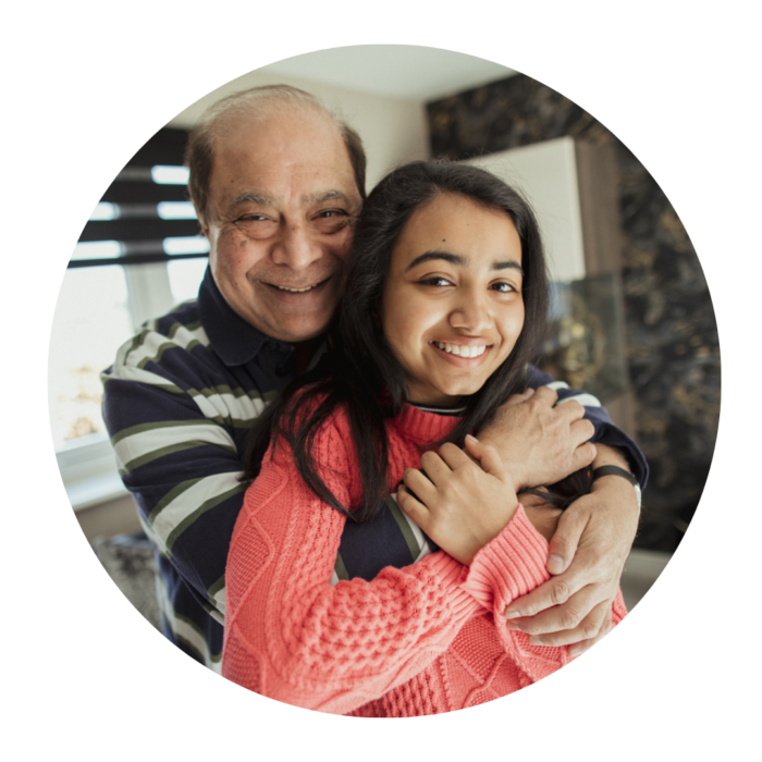 A grandfather hugging his granddaughter as they both look towards the camera and smile for a photo in their house.