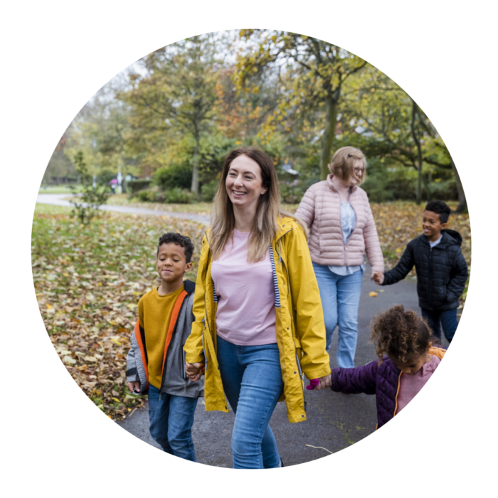 A woman walks through the park smiling and holding hands with two small children. Behind her is another woman holding hands with a child. They are both smiling.