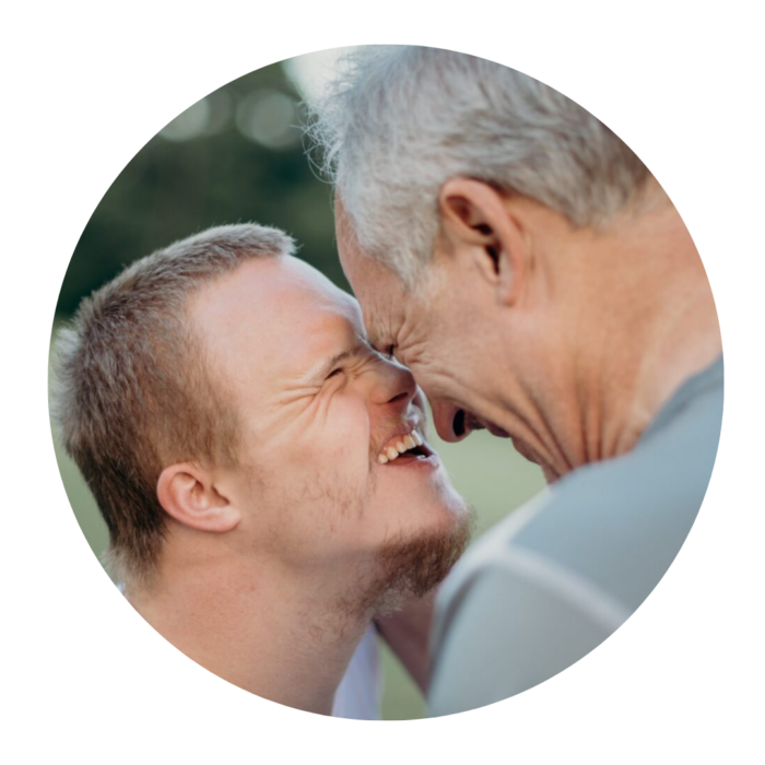 An older man touches his head to the head of a teenage boy with Down's syndrome. They are both laughing.