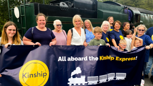 A group of kinship carers and kinship children standing in front of a steam train, holding a banner that says 