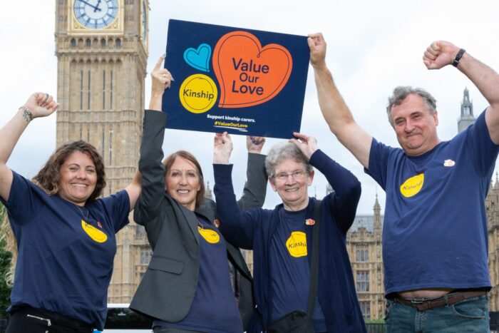 4 kinship carers wearing navy blue Kinship T-shirts with their arms in the air in front of Parliament. Two of them are holding up a Value Our Love placard.