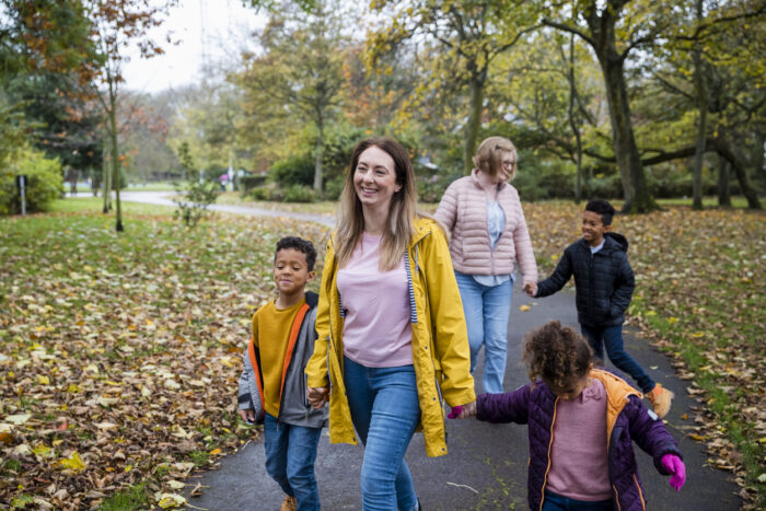 A woman walks through the park smiling and holding hands with two small children. Behind her is another woman holding hands with a child. They are both smiling.