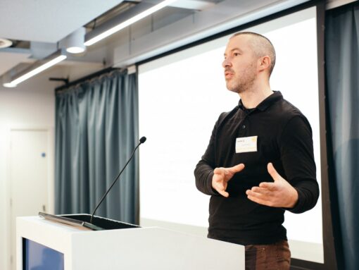 A man with a name tag on stands at a lectern, presenting to a conference.