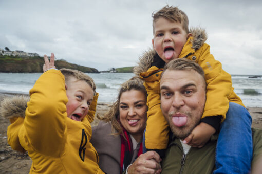 Two adults with two young children on the beach. They are all pulling faces, and one of the children is sitting on one of the adults' shoulders.