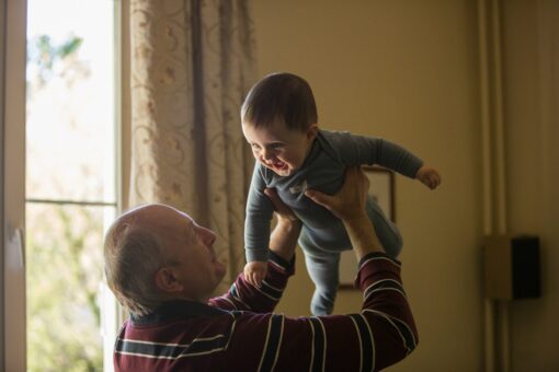 An older man is holding a smiling child high in the air in a living room.