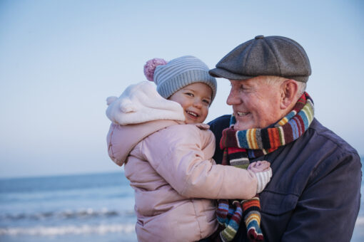 An older man stands holding a small child at the seaside. They are both wrapped up warm and smiling.