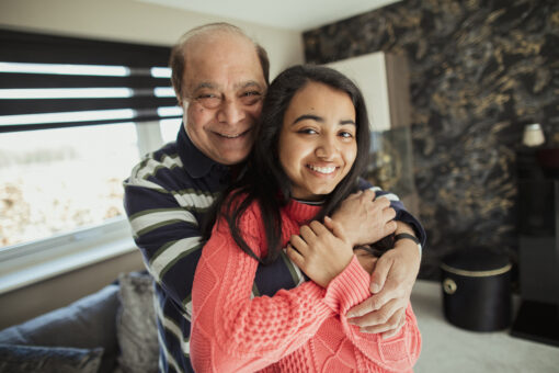 A grandfather hugging his granddaughter as they both look towards the camera and smile for a photo in their house.