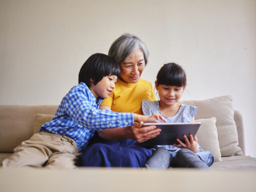 An older woman and two small children sit on a beige sofa and look at a tablet computer. They are all smiling.