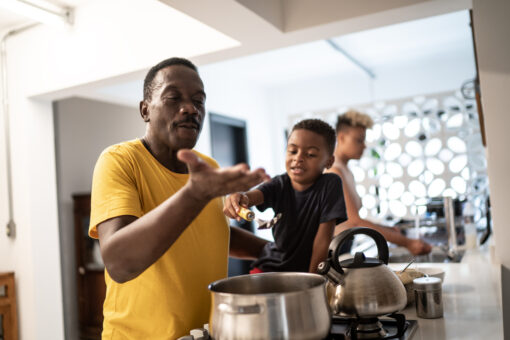 A man is standing over a pot on the stove. A small child is sitting on the counter and handing him a slotted spoon. A second adult is cleaning dishes in the background.