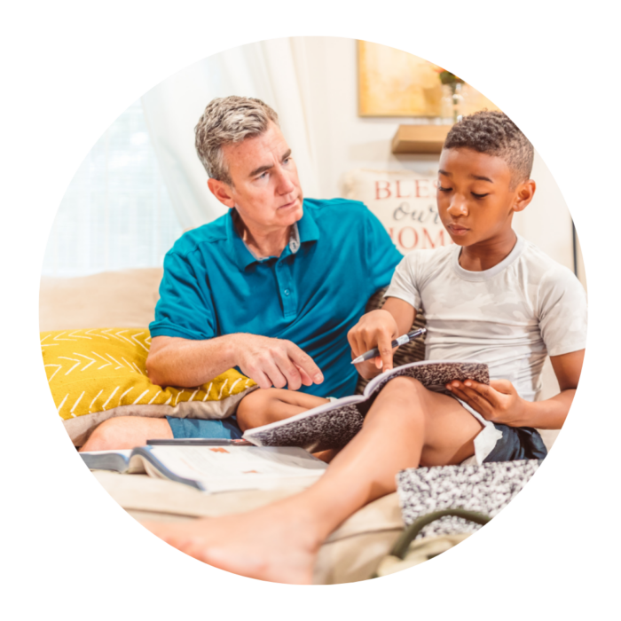 A middle-aged man and a small boy are sitting on a sofa. The man is helping the boy with some homework.