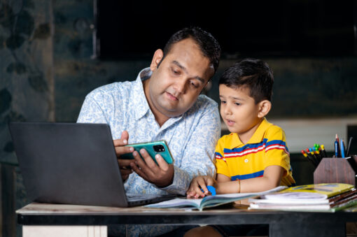 A father showing his son something on a mobile phone. A laptop sits open in front of them.