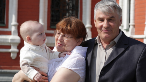 An older man and woman standing outside a house. The woman is holding a baby in her arms.