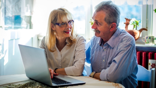 An older couple sit at a table looking at each other, with a laptop in front of them.