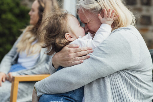 An older woman hugging a young child. They have their noses pressed together and they're both smiling. In the background, another woman sits in a garden chair.