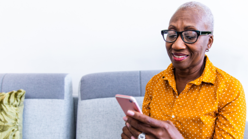 An older woman sits on a grey sofa, smiling as she looks at her mobile phone.