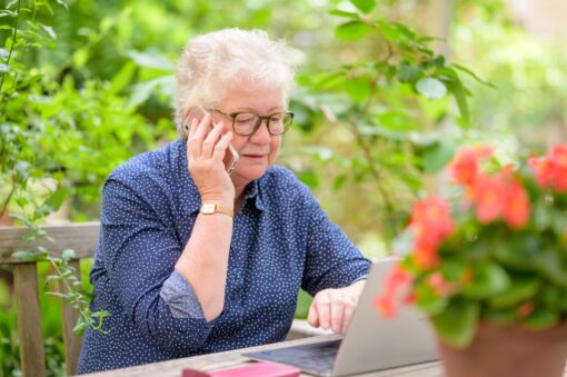 An older woman is sitting outside talking on her phone and looking at a laptop.