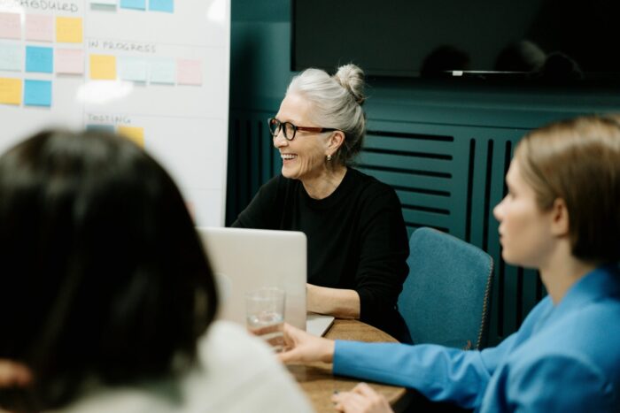 An older woman in a black sweater and glasses sits at her laptop smiling during a work meeting.