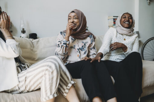 Three women wearing headscarves sitting on a beige sofa, laughing hysterically.