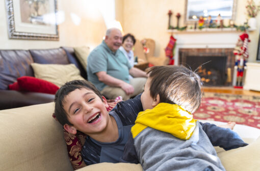 Two small boys playing rough and tumble on a sofa. They're both smiling and laughing. In the background, an older couple sit smiling on a different sofa.