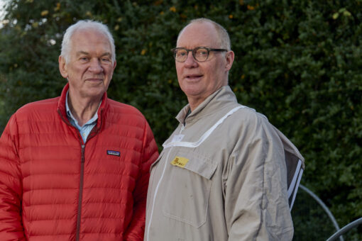 Two older man standing together in front of a hedge.