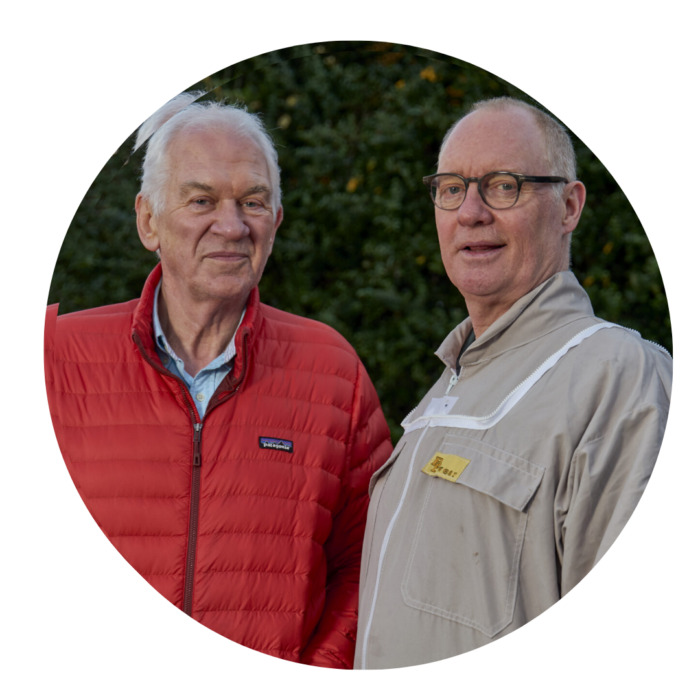 Two older man standing together in front of a hedge.