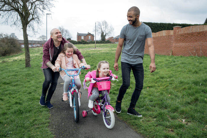 Young male couple taking two small girls out on their bicycles. All four are laughing and smiling.