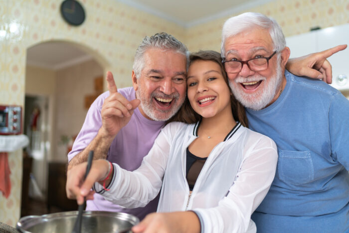 Two older man standing either side of a teenage girl, who is stirring a pot. They are all smiling and looking to camera.