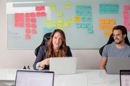 Two people on computers in an office space. Behind them is a whiteboard with lots of post-it notes stuck up.