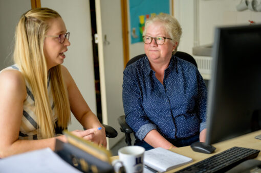 Two women sitting at a computer in an office space chatting.