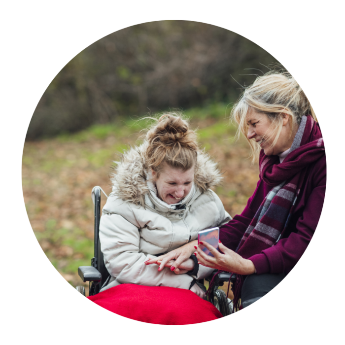 A woman sitting down shows something on her mobile phone to another woman, who is sitting in a wheelchair. They are both wrapped up warm and smiling in the park.