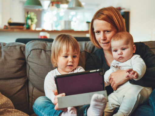 Woman sitting on the sofa with a baby on her lap and another small child next to her. They are all looking at a tablet computer.