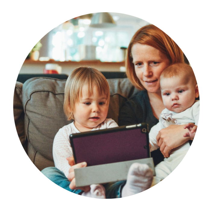 Woman sitting on the sofa with a baby on her lap and another small child next to her. They are all looking at a tablet computer.