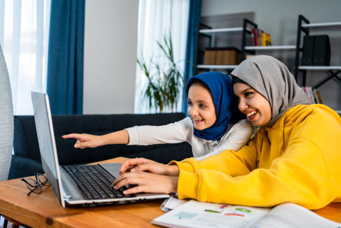 A woman and little girl, both wearing headscarves, look at a laptop. They are both smiling.