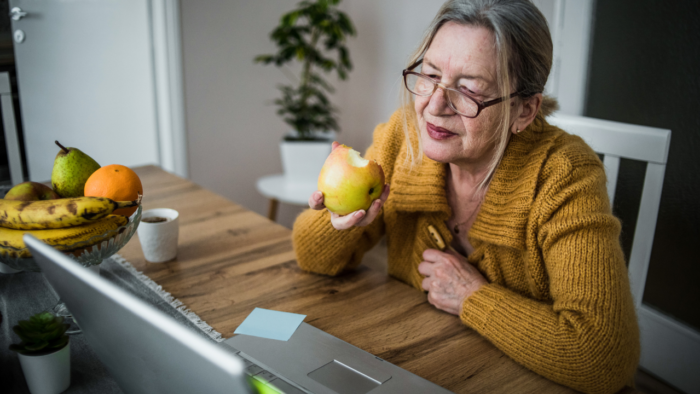 An older woman sitting at the kitchen table eating an apple and looking at her laptop.