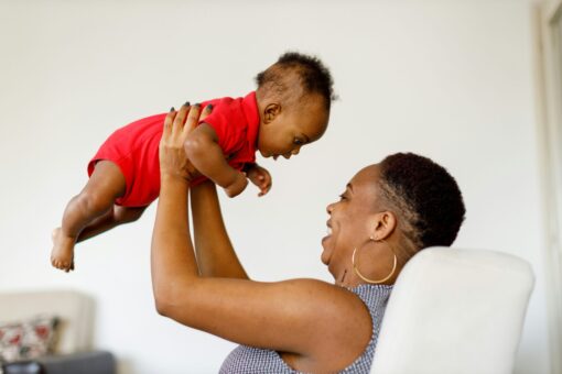 A woman with large hoop earrings is holding a baby in the air and laughing.