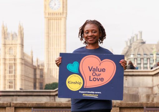 A Kinship campaigner standing across the river from the House of Parliament, holding a placard with the Value Our Love logo.