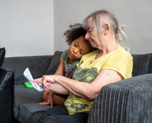 An older woman wearing a yellow T-shirt is reading to a young child, whose head is resting on the woman's shoulder.