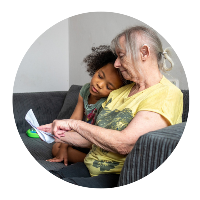 An older woman wearing a yellow T-shirt is reading to a young child, whose head is resting on the woman's shoulder.