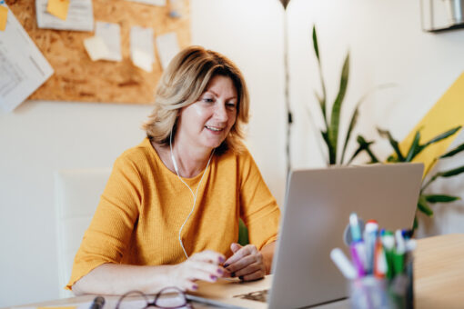 A woman in yellow sits at her laptop. She is wearing earphones and smiling, as if she's on a virtual call.