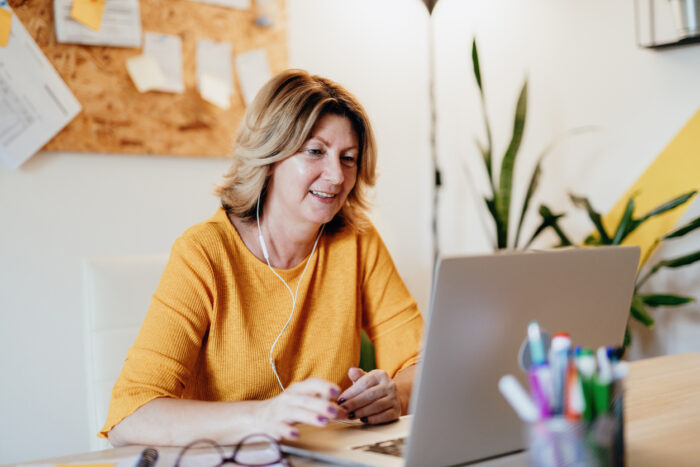 A woman in yellow sits at her laptop. She is wearing earphones and smiling, as if she's on a virtual call.