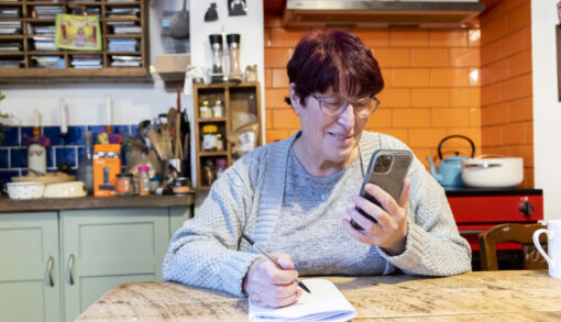 A woman sits at her kitchen table, looking at something on her phone and writing in a notepad.