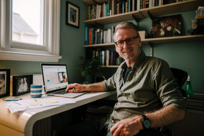 A man sitting at a desk in his study. He has one hand on a laptop, but is turned to look at the camera, smiling.