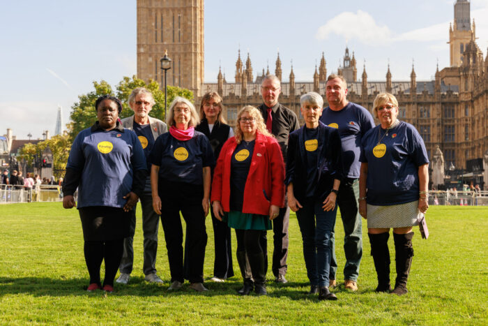 A group of nine kinship carers, all wearing navy blue T-shirts with the Kinship logo on them, stand outside the Houses of Parliament.
