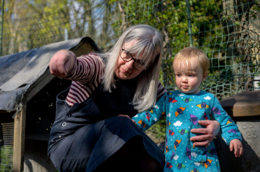 An older woman crouches next to a small child in a garden, beside a chicken coop. The woman is holding chicken feed in her hand.