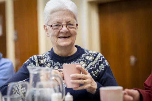 An older woman wearing a navy blue Christmas sweater holds a pink mug and smiles to camera.