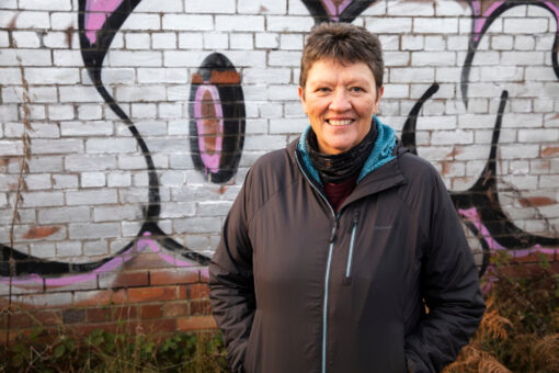 A woman standing in front of a wall covered in graffiti with her hands in her pockets, smiling.