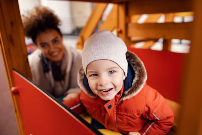 A little boy smiling and having fun in a castle at the playground. Blurred in the background is a smiling adult.
