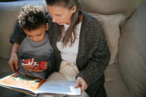 A small child holding a toy truck and reading a book on the sofa with an older woman.